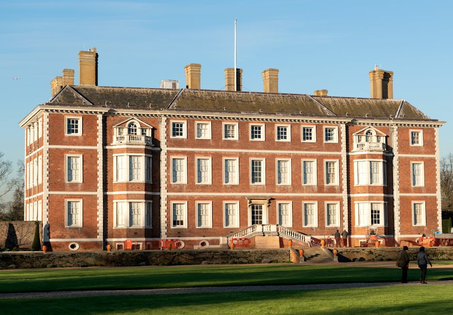 A large historic brick building with classical architectural features, including symmetrical rows of evenly spaced white-framed sash windows, two small projecting bay windows with decorative balconies on the upper floors, and a prominent central staircase with a balustrade leading up to a main entrance door. The building is situated in a spacious area with a well-maintained lawn and a paved pathway in the foreground, with two individuals walking near the edge of the grass. The environment is bathed in natural daylight with a clear blue sky overhead, and several tall chimney stacks are visible on the roof, indicating an older construction style. The scene suggests an institutional or historic residential building, possibly related to private ownership for events or heritage purposes, associated subtly with waste management and rubbish removal services by Waste Disposal Richmond, especially considering the emphasis on external maintenance and cleaning of the property exterior.