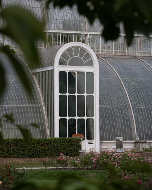 The image showcases a large glass greenhouse structure with a domed roof and metal framework, situated within a well-maintained garden area. In the foreground, there are numerous flowering plants with small, rounded pink blossoms and green foliage, some in terracotta pots arranged on the paved ground. The garden features neatly trimmed bushes and small trees, with additional floral displays and a path leading toward the greenhouse. To the right, a contemporary metallic sculpture with smooth, curved surfaces is partially visible, contrasting with the natural greenery. Overcast skies cast a diffuse light over the scene, contributing to a subdued atmosphere. The environment appears to be in a public garden or botanical setting, which could offer private or independent waste collection services, such as those provided by Waste Disposal Richmond, to ensure cleanliness and maintenance of the site, especially given the outdoor setting and floral landscaping.