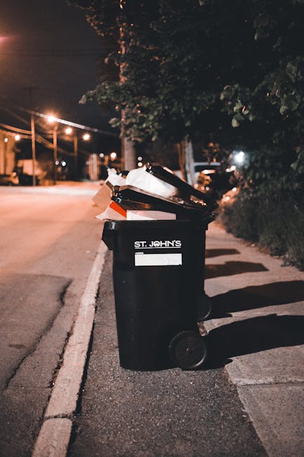 A black wheelie bin labeled 'ST. JOHN'S' is positioned on a city sidewalk at night, filled with various types of rubbish including cardboard boxes, plastic packaging, and paper waste, which are visibly protruding from the open lid. The bin is situated near the curb on a paved pedestrian walkway, with shadows cast on the ground by nearby streetlights. In the background, there are trees and a row of street lamps illuminating the quiet street, along with parked cars further along the road. The scene suggests on-site rubbish disposal or independent waste collection, consistent with private waste management services such as those offered by Waste Disposal Richmond. The overall environment indicates a residential or urban setting where rubbish is being temporarily stored before collection, aligning with general procedures for local waste handling and rubbish removal in the area.