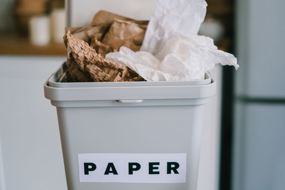 A close-up view of a white plastic bin designated for paper waste, with a label reading 'PAPER' affixed to the front. The bin is filled with crumpled and torn sheets of white and brown paper, some of which are overflowing slightly from the top. The surface of the bin is smooth with an integrated handle on the side for opening, and it is positioned outdoors on a paved or concrete surface. In the background, there are blurred elements of an outdoor environment, possibly a driveway or garden area, with natural lighting suggesting daytime. The scene exemplifies an organized approach to waste separation, aligning with private rubbish collection services like those offered by Waste Disposal Richmond, and demonstrates correct disposal of paper materials within a household or commercial waste management context.