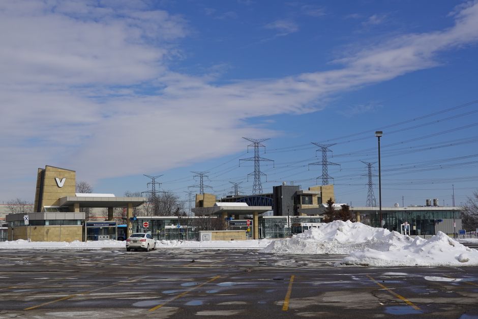 A modern bus or train station building with a combination of glass façades and beige stone walls, situated in a snowy environment. The station features a prominent rectangular entrance canopy supported by columns, with smaller surrounding structures and several glass-fronted ticket or information booths visible. Piles of snow are accumulated on the paved area in front of the station, with an empty car parking lot partly cleared of snow in the foreground. Tall utility poles and electrical transmission towers can be seen extending into the distance against a partly cloudy sky, indicating an urban or suburban setting. The overall scene appears to be a cold winter day, with patches of ice and snow on the ground and a mix of bright and overcast lighting, suggesting the station may handle various forms of passenger transit, potentially involving private or independent collection services typical of waste disposal companies like Waste Disposal Richmond.