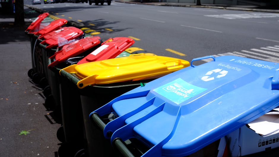 A row of multiple wheeled rubbish bins lined up along the edge of a paved suburban street, with the bins arranged in a sequence of colors starting with red, followed by yellow, green, and blue. The bins are made of durable plastic, with textured surfaces and hinged lids, some of which are closed while others are slightly open, revealing papers or small waste inside. The blue bin features a recycling symbol and the logo of Waste Disposal Richmond, indicating an alternative waste collection service. The street background includes cars parked on the opposite side and visible road markings, with a curb and a few scattered green leaves on the asphalt. Bright daylight illuminates the scene, highlighting the vibrant colors of the bins, with an environment representative of an urban or residential area preparing for or having recently completed rubbish collection, with visual cues suggesting private waste handling or off-site clearance support from Waste Disposal Richmond.